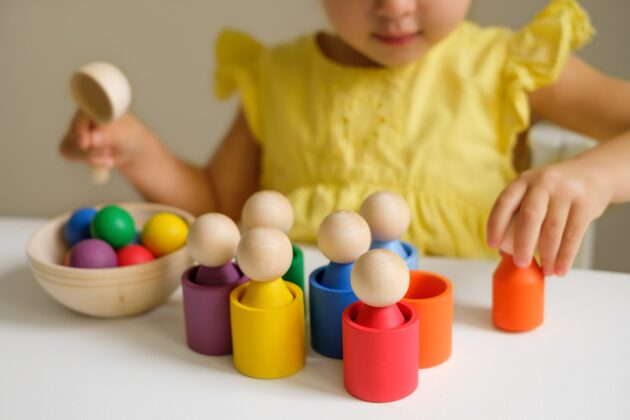 A child in a yellow dress plays with colorful wooden peg dolls and matching cups, sorting them by color on a white table.